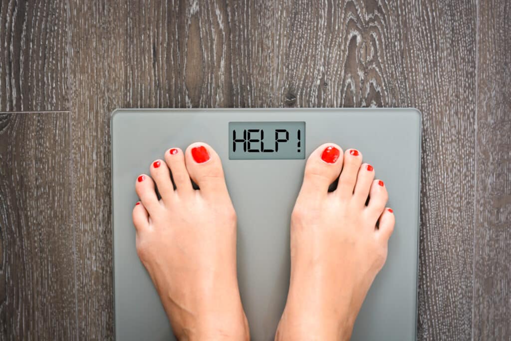 Close up of a woman’s feet standing on a scale and the display says “help” to show she needs help with weight loss.